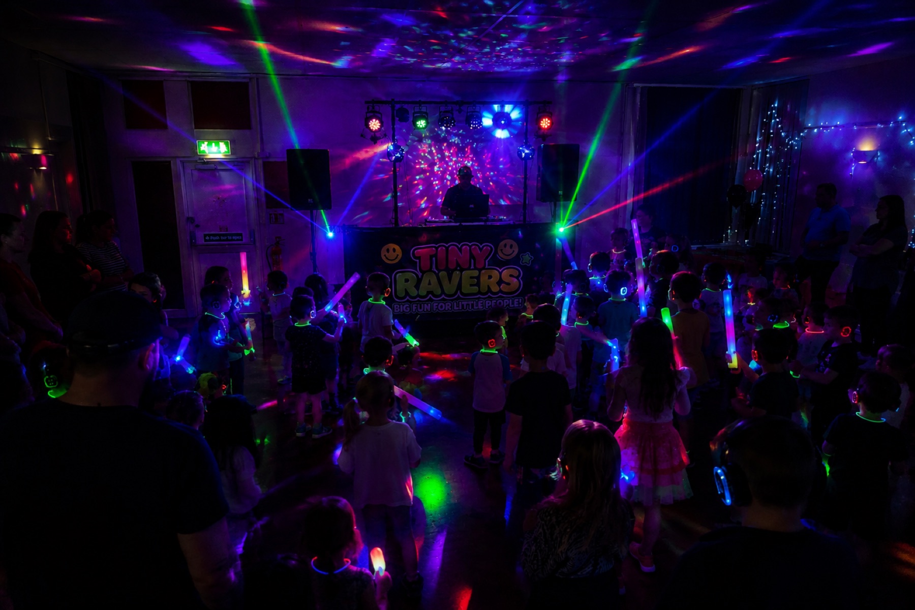 Children dancing with glow sticks in front of the Tiny Ravers DJ booth under lasers and disco lights